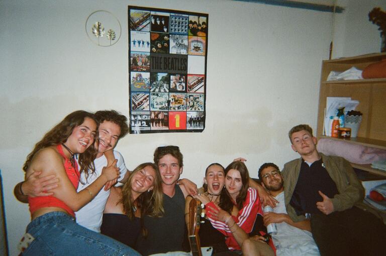 A group of eight friends smile and pose energetically on a couch in a cozy room. A Beatles poster hangs above them, adding a cheerful, nostalgic vibe.
