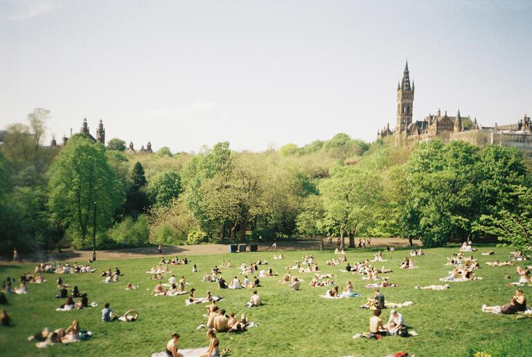 Sunny park scene with people sunbathing on a grass lawn, surrounded by lush green trees. A historic building with a tall tower is visible in the background.