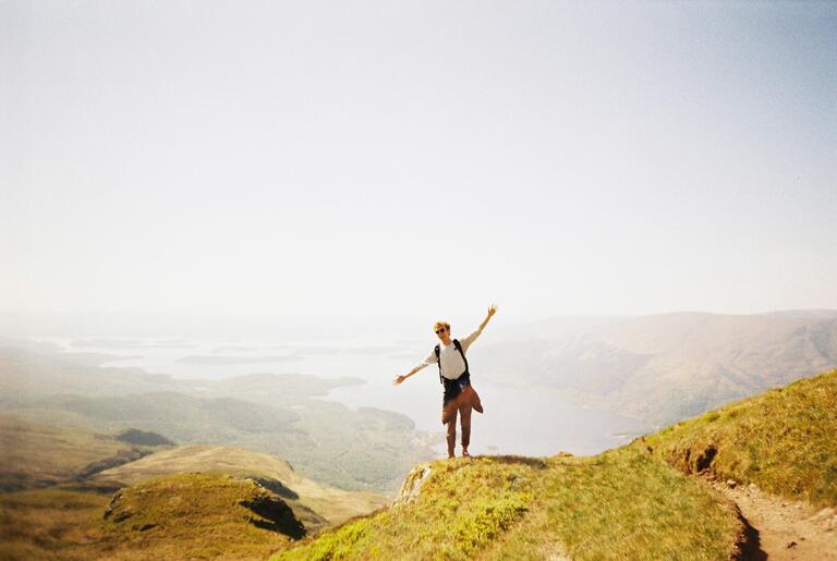 A person stands joyfully on a grassy mountain peak, arms outstretched, overlooking a vast, serene landscape with lakes and distant hills under a clear sky.