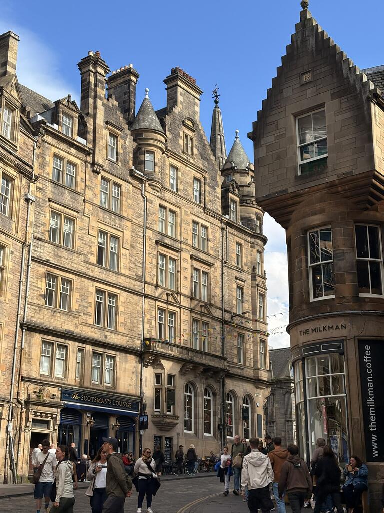 Historic street scene with ornate sandstone buildings under a blue sky. People walk below, adding liveliness to the charming, architectural setting.