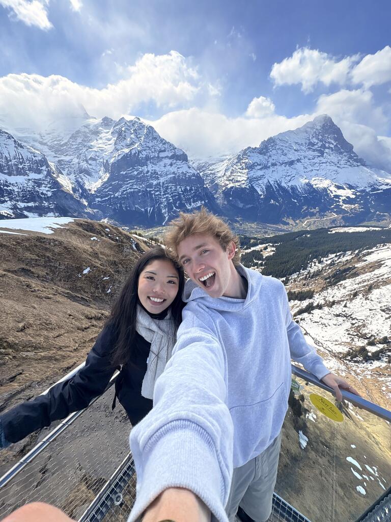 A cheerful duo takes a selfie with snow-capped mountains in the background. Bright sky and dramatic peaks complement their joyful expressions.