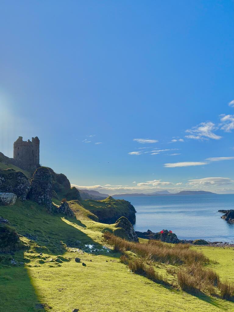 A medieval stone castle towers over a grassy cliff, overlooking a calm blue sea under a bright, clear sky. The scene is serene and picturesque.