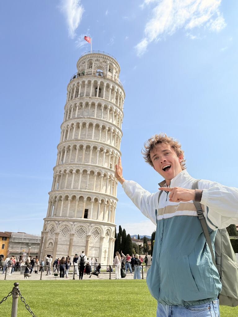 A person playfully poses as if holding up the Leaning Tower of Pisa. They smile excitedly on a sunny day, with tourists milling around in the background.