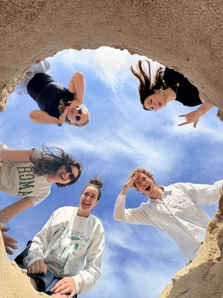 Five people smiling and posing while looking down into a sand pit, with a blue sky above. The scene conveys a playful, joyful atmosphere.