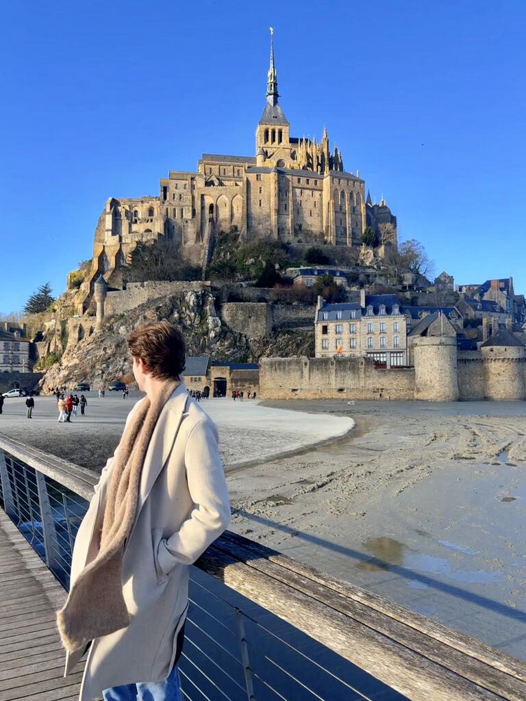 Person in a coat gazes at Mont Saint-Michel, France, under a clear blue sky. The historic abbey stands majestically on a rocky hilltop.