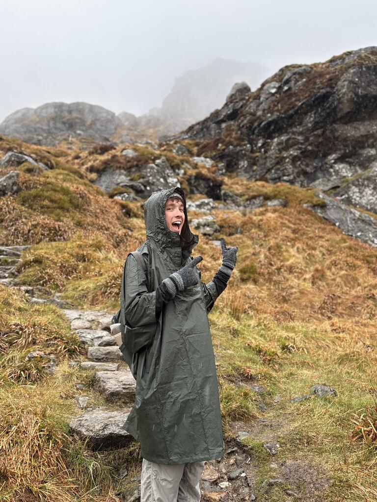 A person in a rain poncho smiles and gestures with excitement while hiking on a misty, rocky trail. The terrain is covered in wet, orangey-brown grass.
