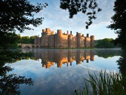 A view of Herstmonceux Castle across the moat framed by trees with a reflection of the castle in the water