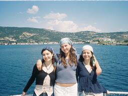 Three smiling women stand arm-in-arm on a boat, wearing headscarves against a backdrop of blue water and hilly landscape under a sunny sky.