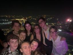 A group of nine friends smiling at a nighttime rooftop gathering. City lights twinkle in the background, creating a festive and joyful atmosphere.