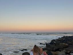 Mya and friend sit on rocks by the ocean, embraced, gazing at a serene sunset. The sky transitions from soft orange to blue, creating a peaceful mood.
