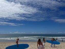 A group of women carrying blue surfboards walks on a sandy beach towards the ocean. The sky is partly cloudy with a serene and relaxed vibe.
