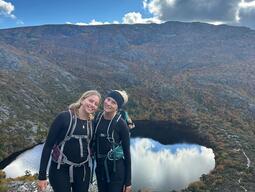 Mya and friend smile on a rocky mountain trail with a vibrant landscape of a lake and autumn-colored forest under a partly cloudy sky in the background.
