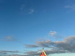 A boat sails on a calm ocean under a clear blue sky with scattered clouds. In the foreground, the Australian flag waves, and a green coastline is visible in the background.