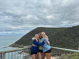 Three friends hug on a wooden deck overlooking a scenic coastal view with lush green hills and a cloudy sky, conveying joy and togetherness.