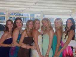 Seven women stand closely together in colorful dresses, smiling warmly. Background shows a bright, covered outdoor area with cityscape glimpses.