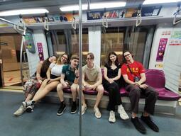 A group of six friends sitting closely on a subway train's purple seats, smiling and casually posing. The atmosphere is lively and relaxed.