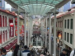 Bustling street market scene with diverse crowds between historic buildings adorned with signs. A glass roof overhead creates an urban, vibrant atmosphere.