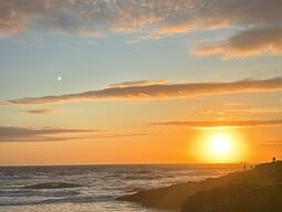 Sunset over a rocky coastline with gentle waves and scattered clouds in the sky. The warm glow creates a serene and tranquil atmosphere.