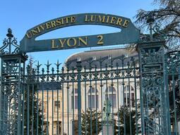 Ornate iron gate of Université Lumière Lyon 2, with a grand building and statue visible behind, set in a peaceful garden setting.