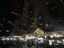 A bustling nighttime market scene, filled with people dining under a canopy of bright lights, is framed by towering, illuminated skyscrapers above.