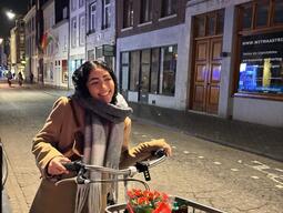 A smiling woman in a tan coat and scarf stands beside her bicycle on a cobblestone street at night. The bike basket holds flowers and snacks.