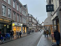 A quaint European street adorned with holiday decorations, including star-shaped lights. People walk past shops on a cobblestone street, creating a cozy atmosphere.