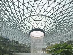 A large indoor waterfall cascades from a geometric glass-domed ceiling into a circular pool, surrounded by lush greenery and people on a walkway.