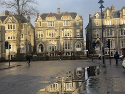 Historic stone buildings with gabled roofs line a wet street, reflecting in puddles. A few pedestrians and cyclists move under a cloudy sky.