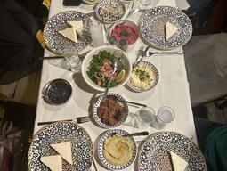 A table set for dinner featuring patterned plates with slices of cheese, colorful salads, dips, and bread. Glasses of clear beverages and utensils are neatly arranged, creating a cozy and inviting atmosphere.
