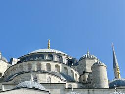 Majestic view of a historic mosque with multiple domes and a tall minaret under a clear blue sky, featuring intricate Arabic calligraphy on the facade.