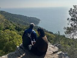 Two people sit on a rock cliff, overlooking a vast, sunlit ocean. The coastline is lush with green trees, creating a serene, peaceful atmosphere.