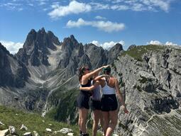 Three people embrace on a grassy mountain ledge, overlooking dramatic jagged peaks. The sky is clear with scattered clouds, conveying adventure and camaraderie.