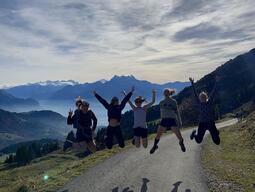 Alyssa and friends jumping in the air with arms outstretched, with mountains and water in the background.