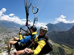 Alyssa and a man paragliding with a landscape of mountains and a sprawling city below. They wear helmets and colorful jackets, exuding excitement and adventure.