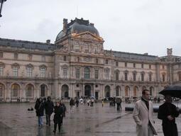 Wet cobblestones reflect a cloudy afternoon in front of the Louvre Museum, Paris. People walk with umbrellas, creating a bustling yet serene atmosphere.