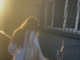 Woman in long skirt and sweater gently pets a cat on a sunlit sidewalk. Warm, golden light from the setting sun creates a serene atmosphere.
