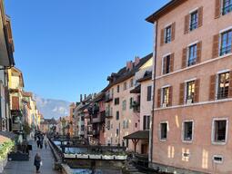 Charming street in France, lined with colorful buildings along a narrow canal. People stroll under a clear blue sky, with mountains in view.