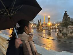 Aidenne with with an umbrella stands on a Parisian bridge at dusk, gazing towards the illuminated Eiffel Tower. The scene is calm and reflective.