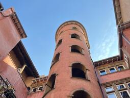 Tall, pink cylindrical tower with arched openings, surrounded by older buildings. Clear blue sky above, conveying a historic and serene atmosphere.