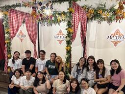 A group of sixteen people smiles for a photo inside a decorated restaurant with floral arches and red checkered curtains. The mood is cheerful and friendly.