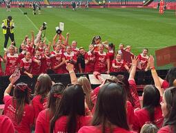 A large group of excited people in red shirts with "#TeamCardiff" printed on the back celebrate together in a stadium, displaying team spirit and unity.