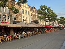 Street view of a café-lined avenue, featuring tables with patrons under awnings. Historic buildings and large trees create a relaxed, inviting atmosphere.