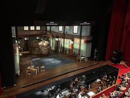 A theater stage set with a large rock and sparse furnishings, viewed from above. An orchestra is in the pit, and an audience fills red seats, creating an anticipatory atmosphere.