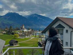 A person in a scarf and jacket stands on a balcony, gazing at a scenic view of mountains, a castle, and cloudy skies. The tone is serene and contemplative.