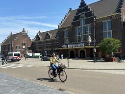A person rides a bicycle in front of Maastricht train station, a historic brick building with stepped gables under a clear blue sky. A peaceful, sunny day.
