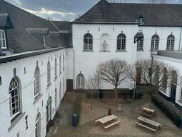 View of the UCM terrace, with white brick buildings, arched windows, bare trees, and wooden picnic tables. A cloudy sky adds a serene atmosphere.
