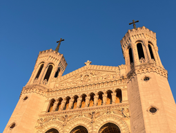 Ornate cathedral façade with two tall towers under a bright blue sky. Warm sunlight highlights detailed carvings and arched windows.