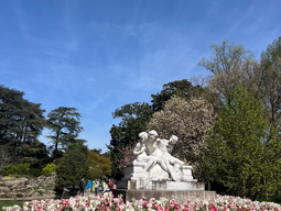 A stone statue of three figures sits among blooming pink and white tulips under a clear blue sky, surrounded by lush green trees, conveying serenity.
