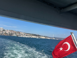 A Turkish flag flutters at the rear of a boat, leaving a foamy trail on the blue waters. Cityscape and clear skies are visible along the distant shore.