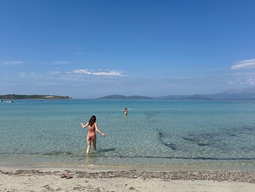 A woman wades into clear, turquoise water at a serene beach under a bright blue sky. Distant islands and scattered clouds create a tranquil scene.